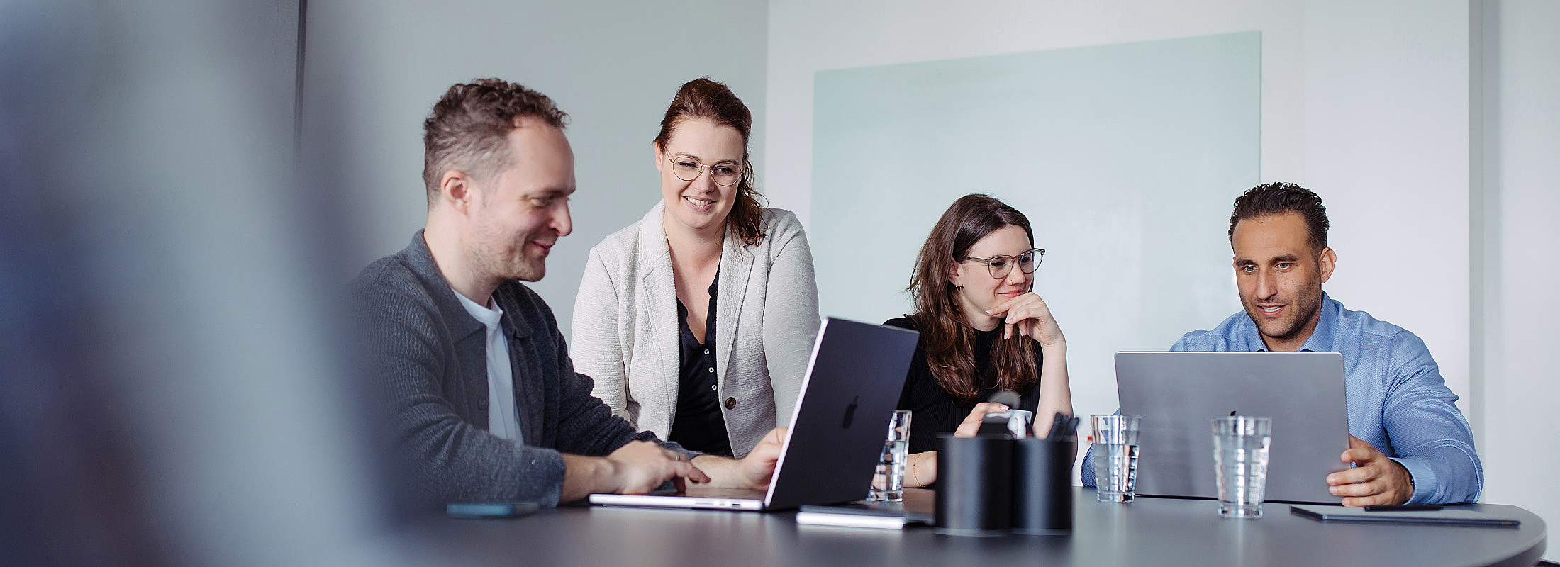 Vier Personen sitzen gemeinsam an einem Tisch im Besprechungsraum, lächeln und arbeiten an Laptops.