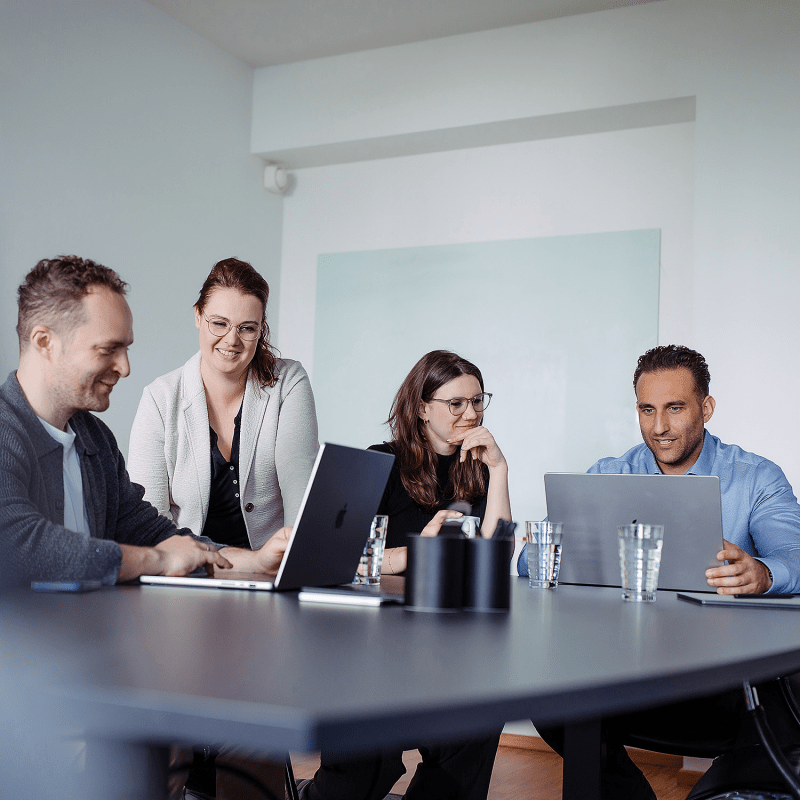 Vier Personen sitzen an einem Konferenztisch mit Laptops und besprechen gemeinsam ein Projekt in einer modernen B&uuml;roumgebung.