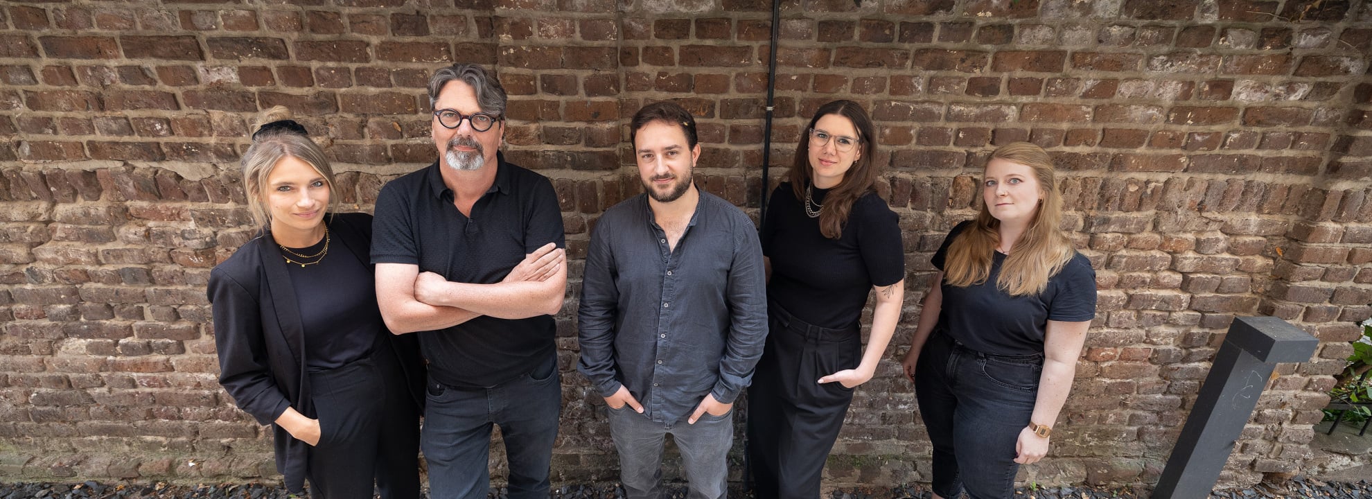 F&uuml;nfk&ouml;pfiges Team in dunkler Kleidung vor begr&uuml;nter Backsteinwand